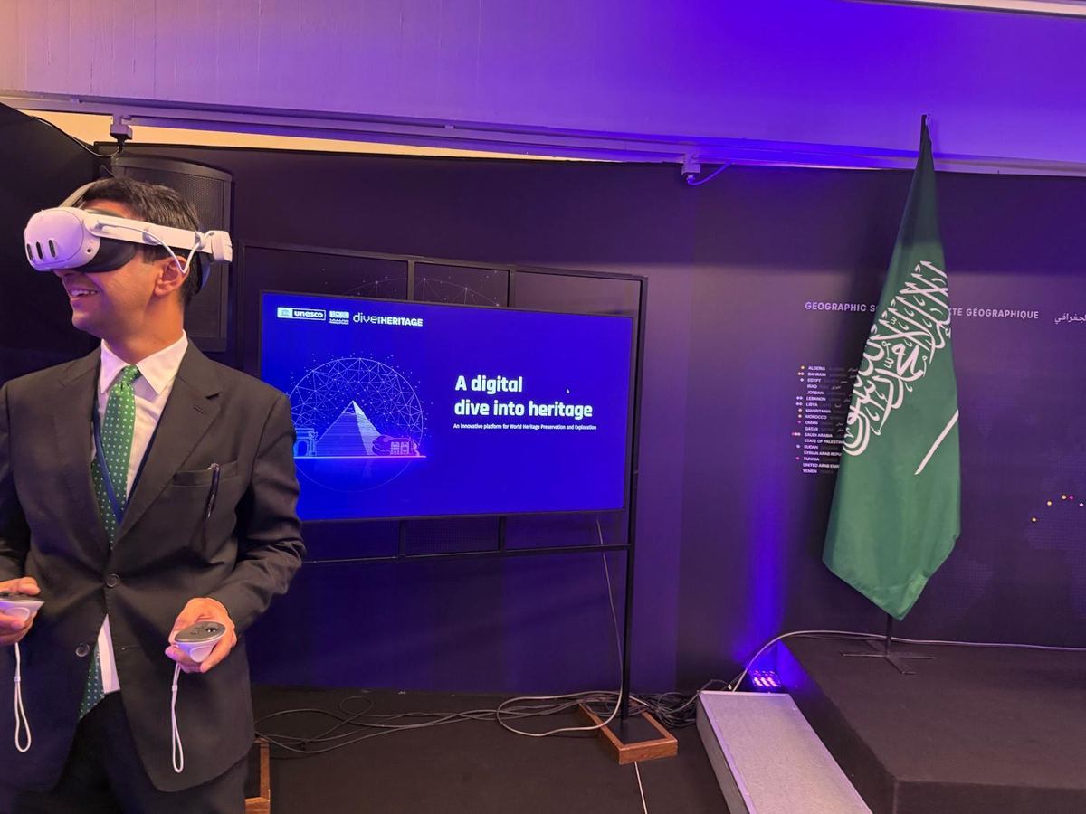 Un visiteur en costume, casque de réalité virtuelle sur la tête et manettes en main, devant l’écran « A digital dive into heritage »