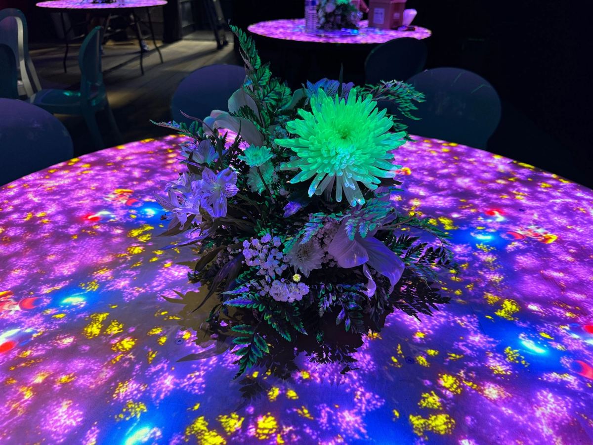 Table ronde du Pavillon France avec un bouquet de fleurs au centre, la nappe recouverte d’une projection violette, jaune et bleue