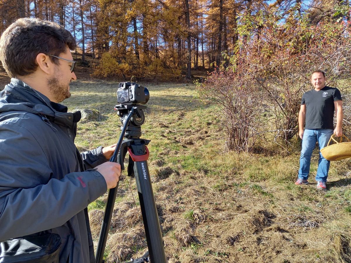 Un cadreur derrière sa caméra sur trépied filme un homme qui tient un panier en osier, devant des mélèzes dorés