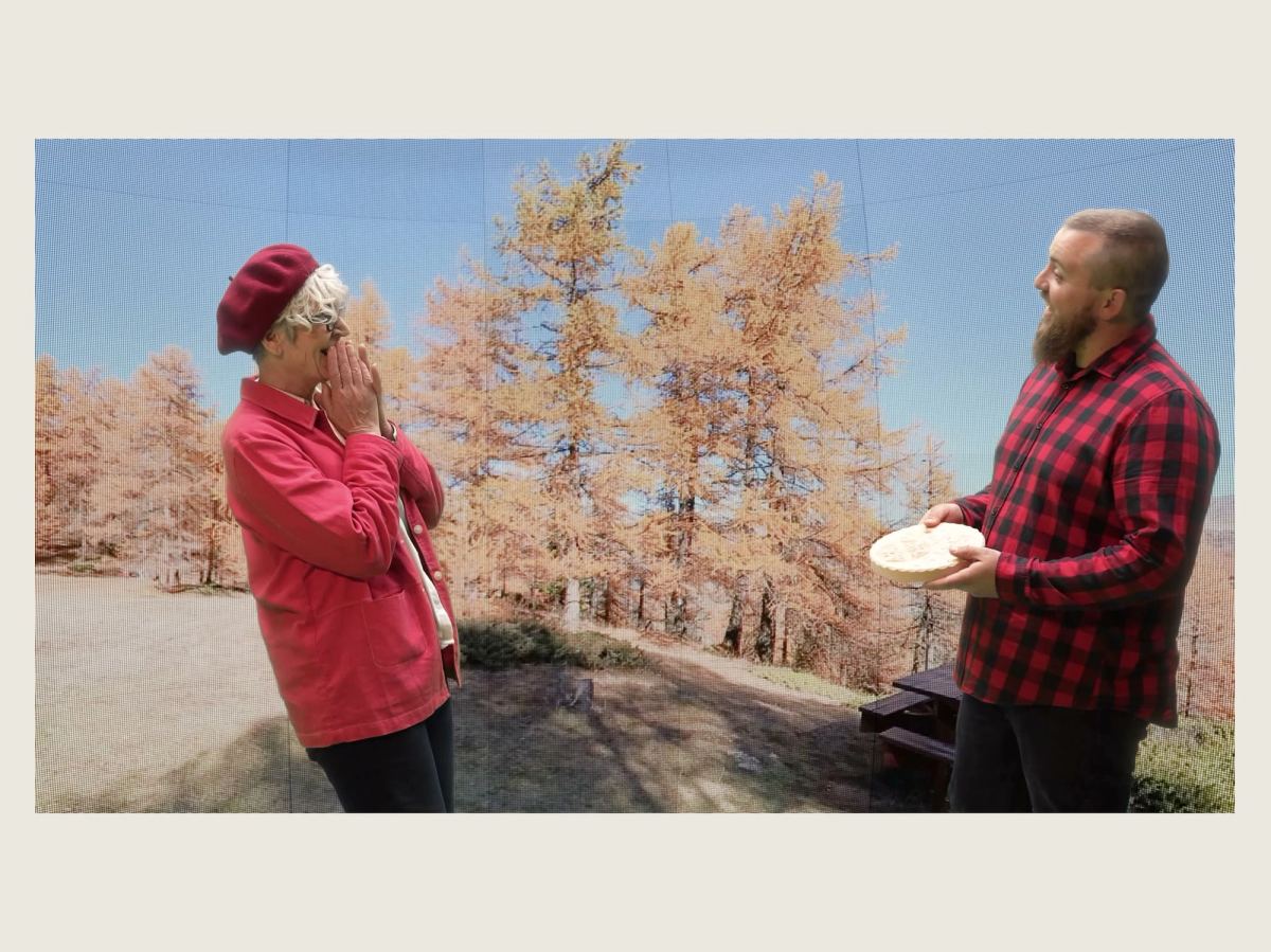 Image du film : une femme en béret rouge, les mains devant la bouche, face à un homme barbu en chemise à carreaux qui lui tend une pièce de bois sculptée