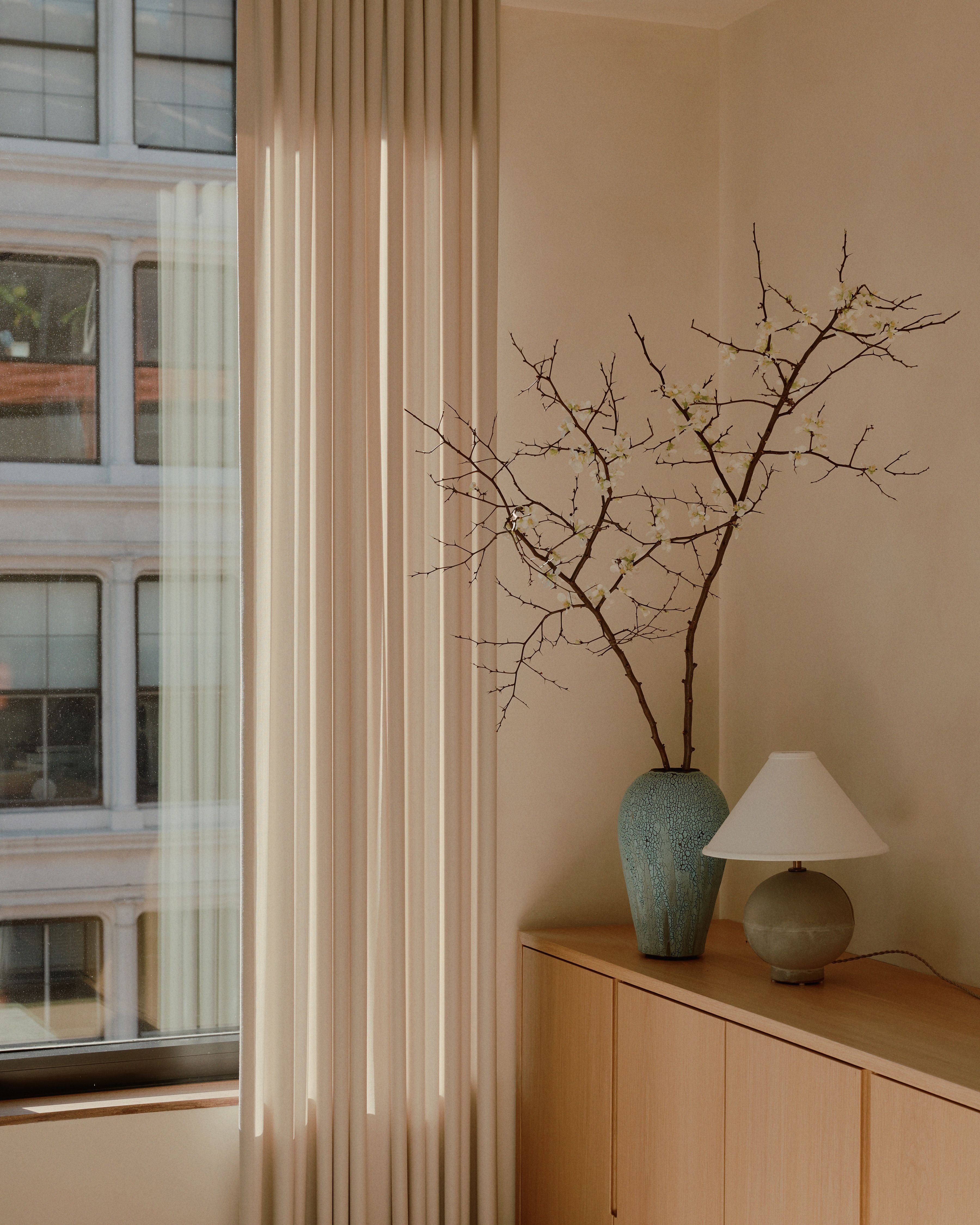 A small lamp and blue vase holding white flowers resting on a credenza next an airy, light filled window.