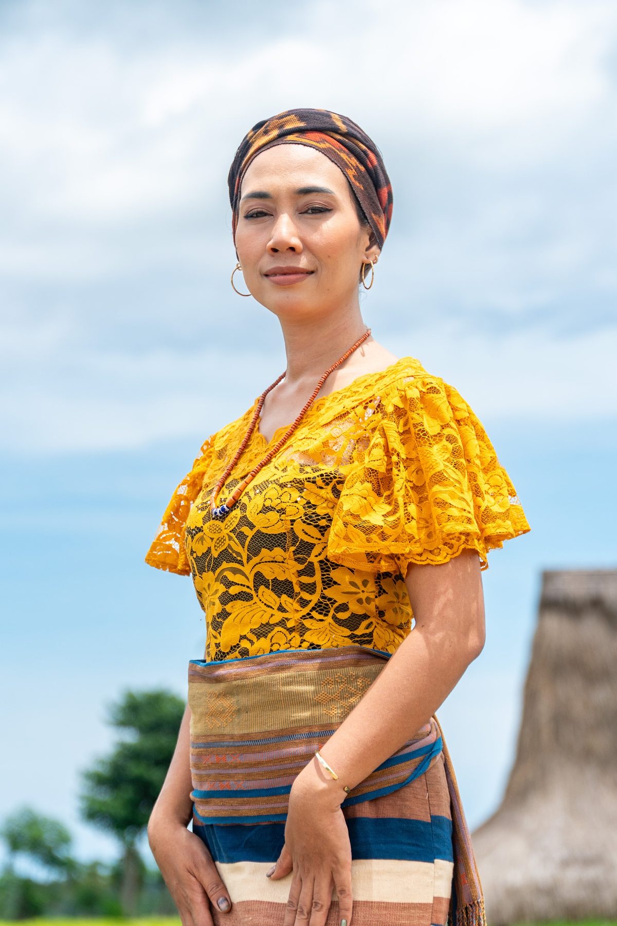 Nensi in a yellow blouse and Ikat beside a traditional thatched roof