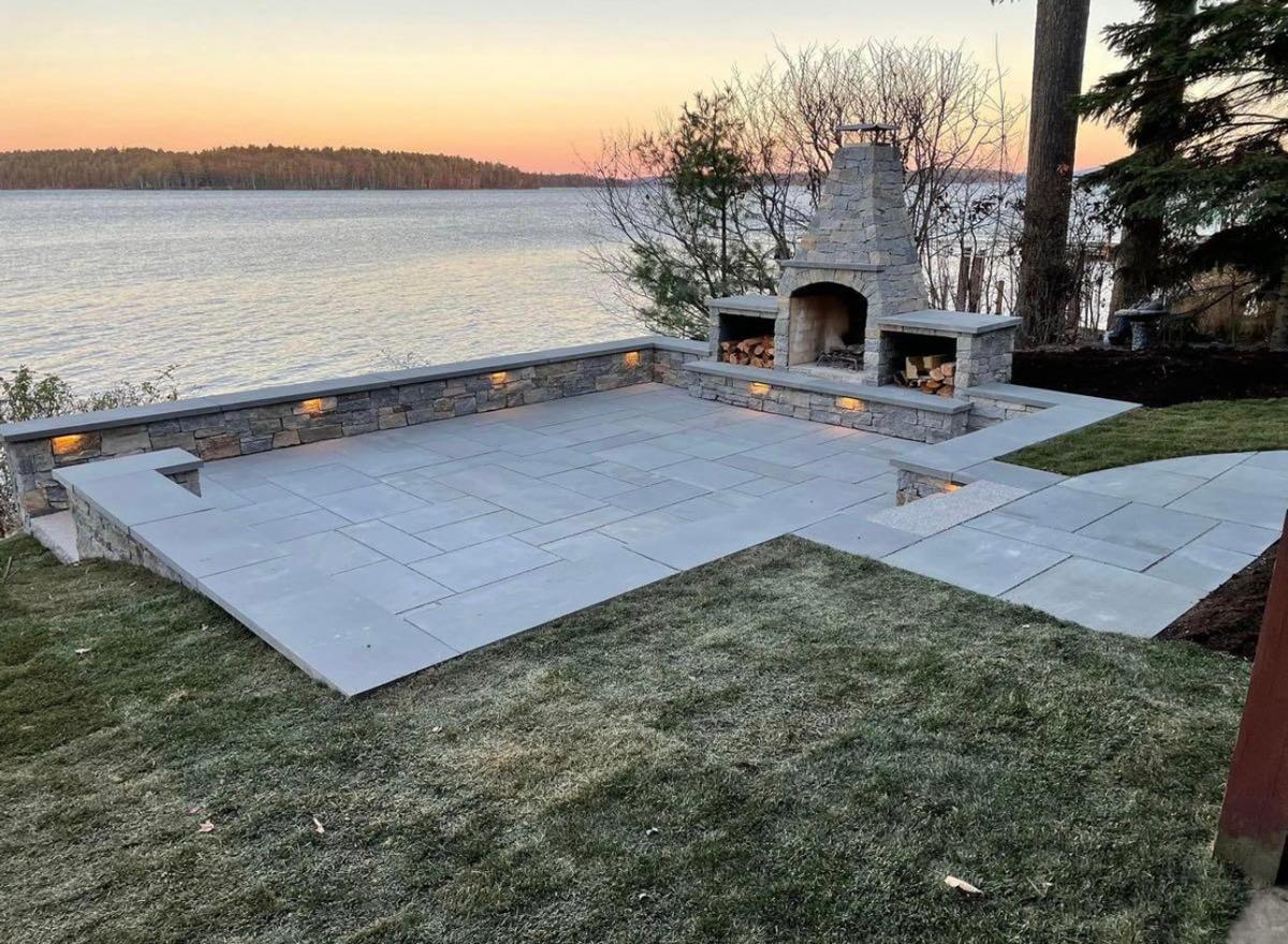 Lakeside patio with custom stone fireplace, bluestone pavers, and built-in wall lighting at sunset