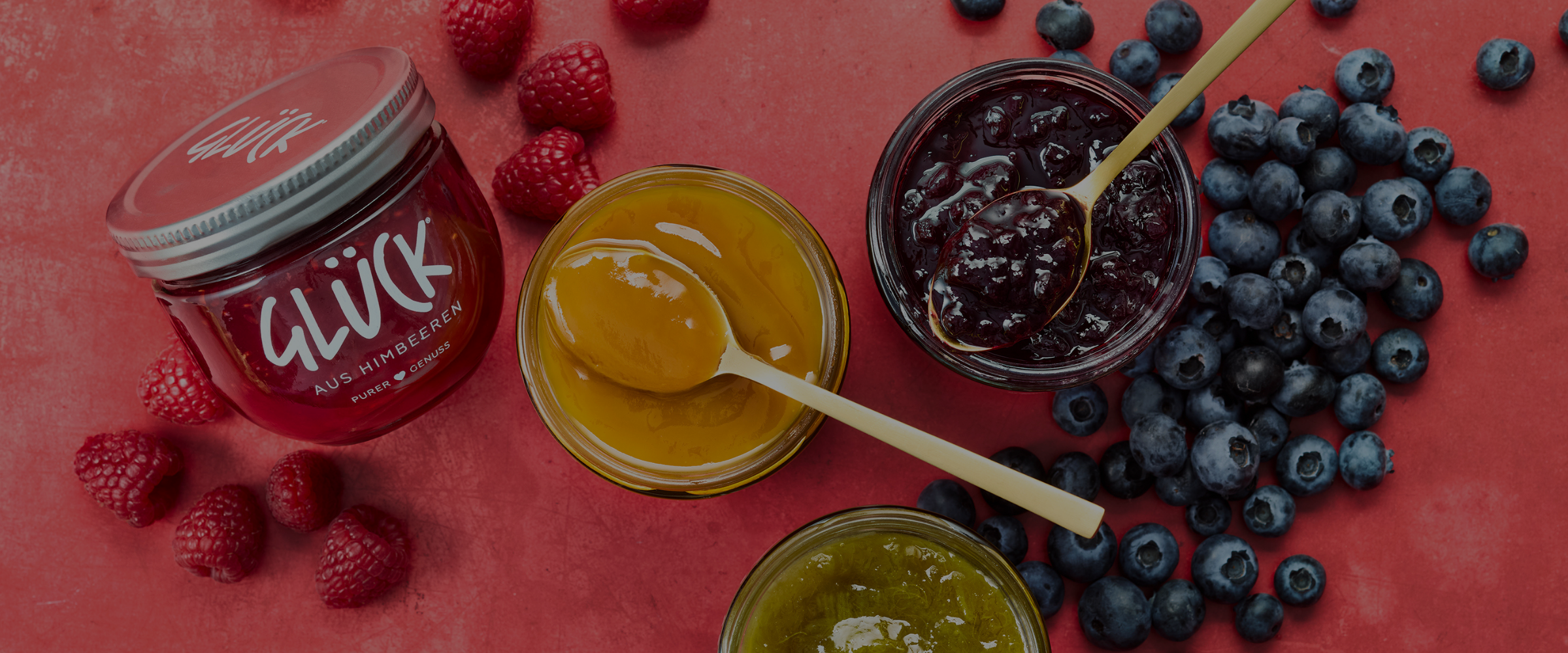 Drei offene Gläser GLÜCK Fruchtaufstriche stehen auf rotem Untergrund. Das untere Glas ist zur Hälfte abgeschnitten. Die beiden offenen Gläser in der Mitte haben je einen Löffel mit Marmelade darauf dort liegen. Links steht ein volles, geschlossenes Glas GLÜCK aus Himbeeren. Im Hintergrund liegen einige Himbeeren und Heidelbeeren.