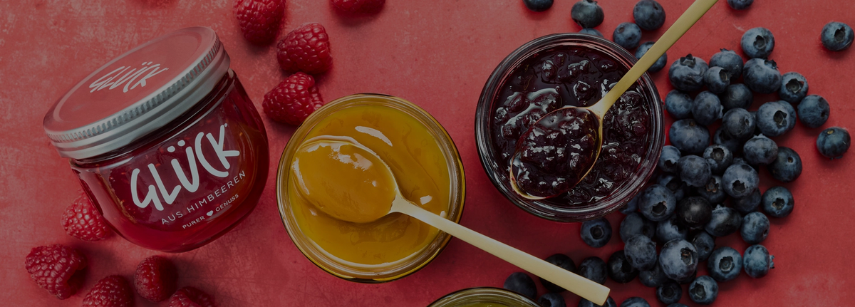 Drei offene Gläser GLÜCK Fruchtaufstriche stehen auf rotem Untergrund. Das untere Glas ist zur Hälfte abgeschnitten. Die beiden offenen Gläser in der Mitte haben je einen Löffel mit Marmelade darauf dort liegen. Links steht ein volles, geschlossenes Glas GLÜCK aus Himbeeren. Im Hintergrund liegen einige Himbeeren und Heidelbeeren.