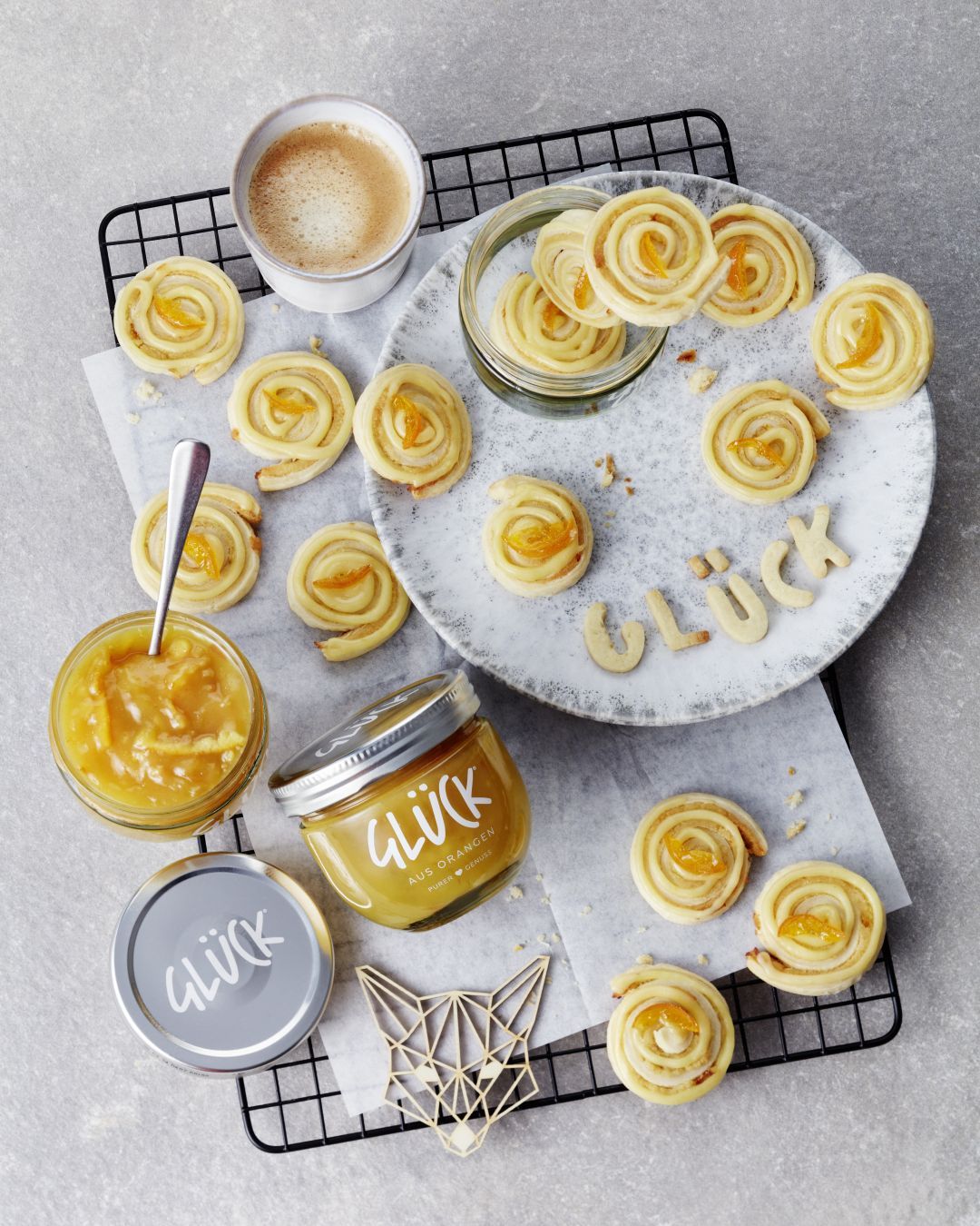 Orangen-Marzipan-Schnecken auf einem Gitter, daneben ein Glas GLÜCK aus Orangen.