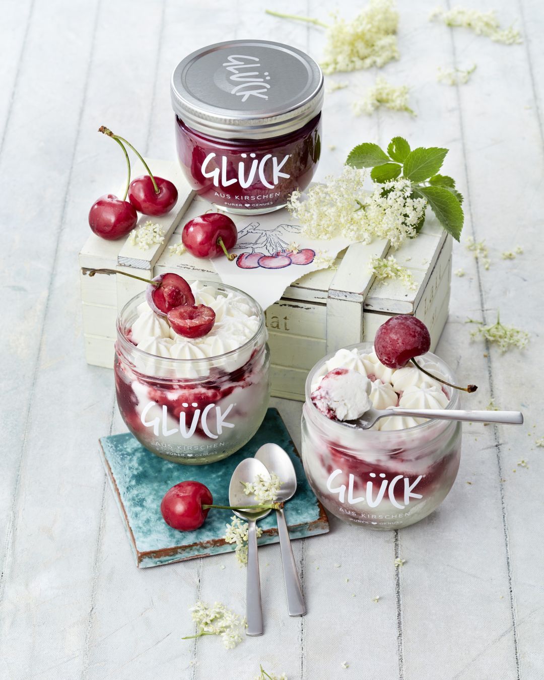 Semifreddo mit Kirschen und Holunderblüten im GLÜCK Glas, garniert mit Sahne und frischen Kirschen, daneben ein Glas GLÜCK aus Kirschen. Daneben weitere Kirschen, Löffel, Holunderblüten und ein Glas GLÜCK aus Kirschen.