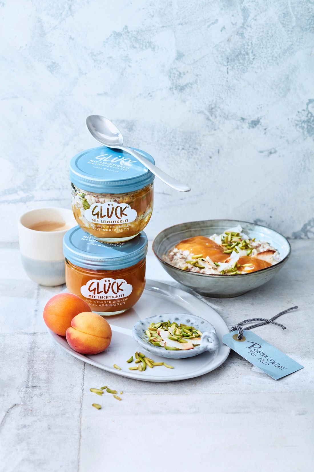Frühstücksporridge mit Aprikosen-Topping im GLÜCK Glas. Darunter steht ein Glas GLÜCK mit Leichtigkeit aus Aprikosen. Als Dekoration daneben Aprikosen, Kerne und eine volle Schüssel des Porridge.