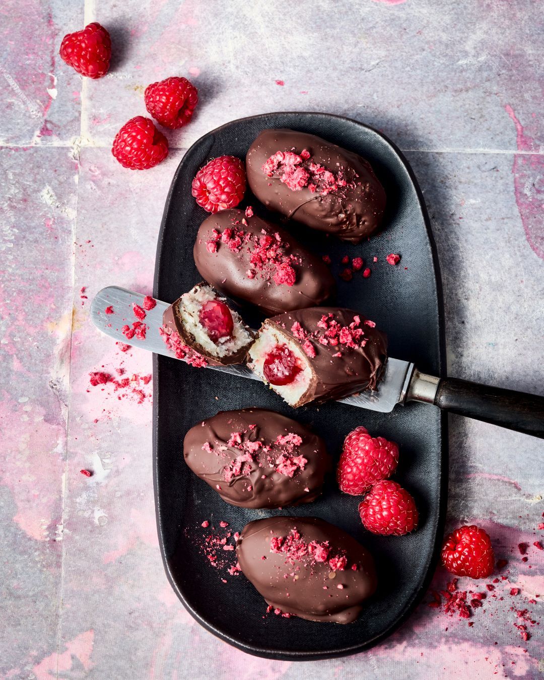Himbeeren-Kokosbars gefüllt mit GLÜCK aus Himbeeren auf einem schwarzen Tablett. Ein Kokosbar ist angeschnitten und auf einem Tortenanheber drapiert.