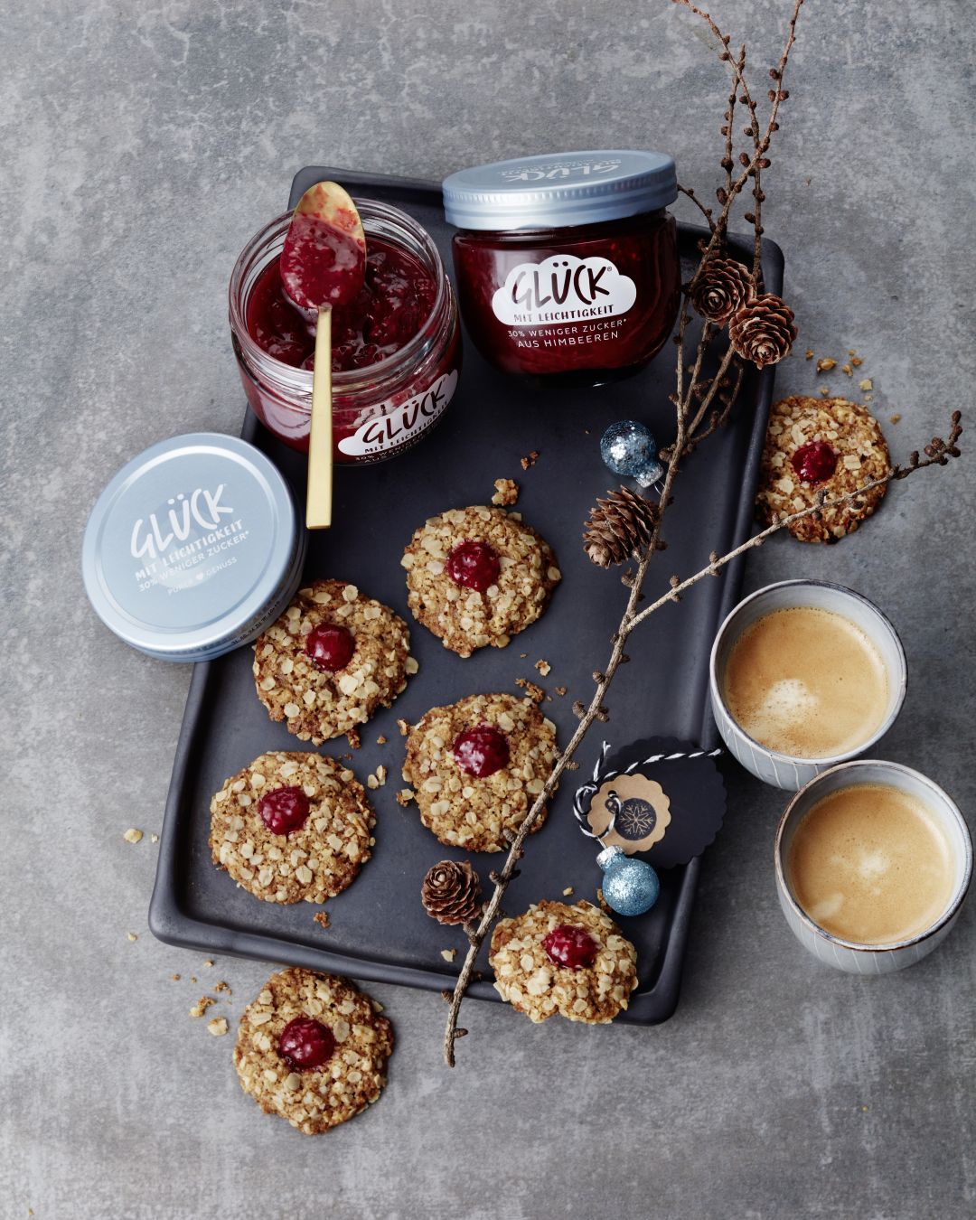Haferflockenplätzchen mit GLÜCK mit Leichtigkeit aus Himbeeren. Daneben ein Glas GLÜCK mit Leichtigkeit aus Himbeeren. Zur Deko Kaffeebecher und ein Ast.