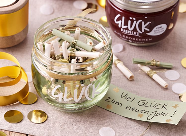 Ein leeres GLÜCK Glas, gefüllt mit kleinen aufgerollten Zetteln mit goldenen Bändchen. Drum herum liegt Silvesterdekoration und ein Zettel mit der Aufschrift "Viel GLÜCK zum neuen Jahr!"