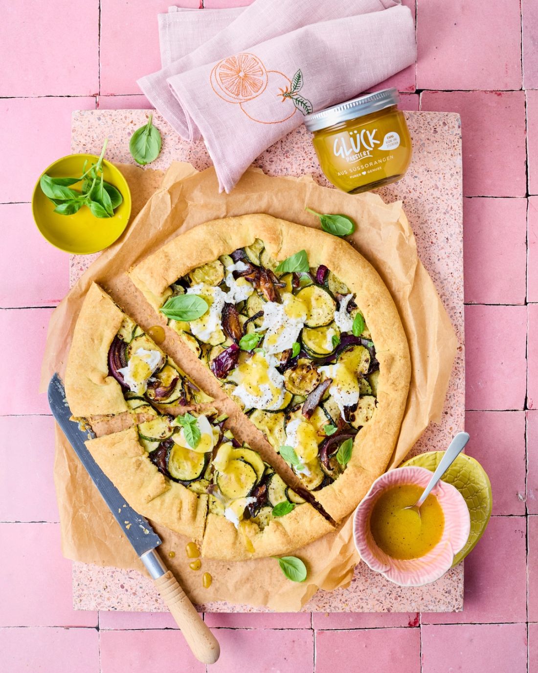 Zucchini-Galette mit Burrata und Orangen-Balsamico, angerichtet auf rosa Fliesen mit Basilikum und einem Glas GLÜCK passiert aus Süßorangen