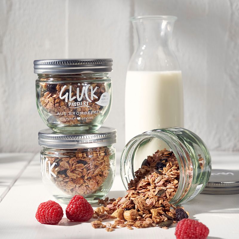 Granola im GLÜCK Glas, daneben Milch und Himbeeren. Ein Glas Granola ist halb ausgekippt und das Granola davor verstreut.