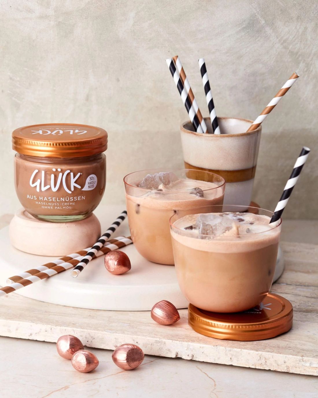Iced Hazelnut Coffee in Gläsern mit Strohhalmen. Daneben ein Glas GLÜCK aus Haselnüssen.