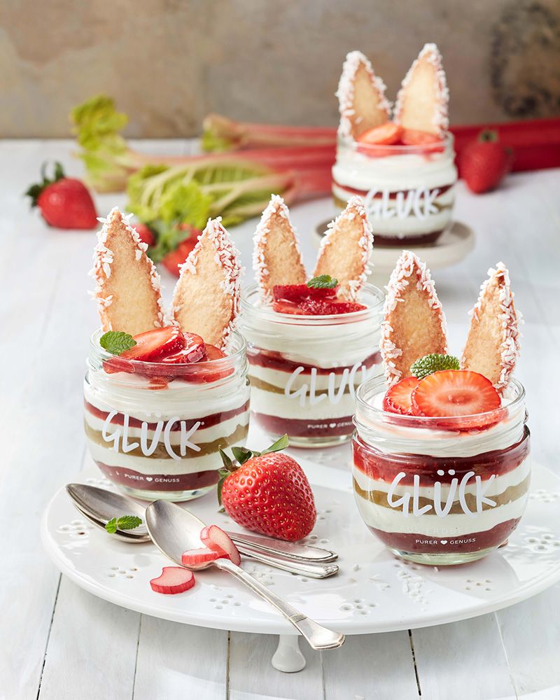 Geschichtetes Erdbeer-Rhabarber-Osterdessert mit Hasenohren und Erdbeeren im GLÜCK Glas. Daneben Erdbeeren und Löffel. Auf dem Teller stehen drei Dessert-Gläser.