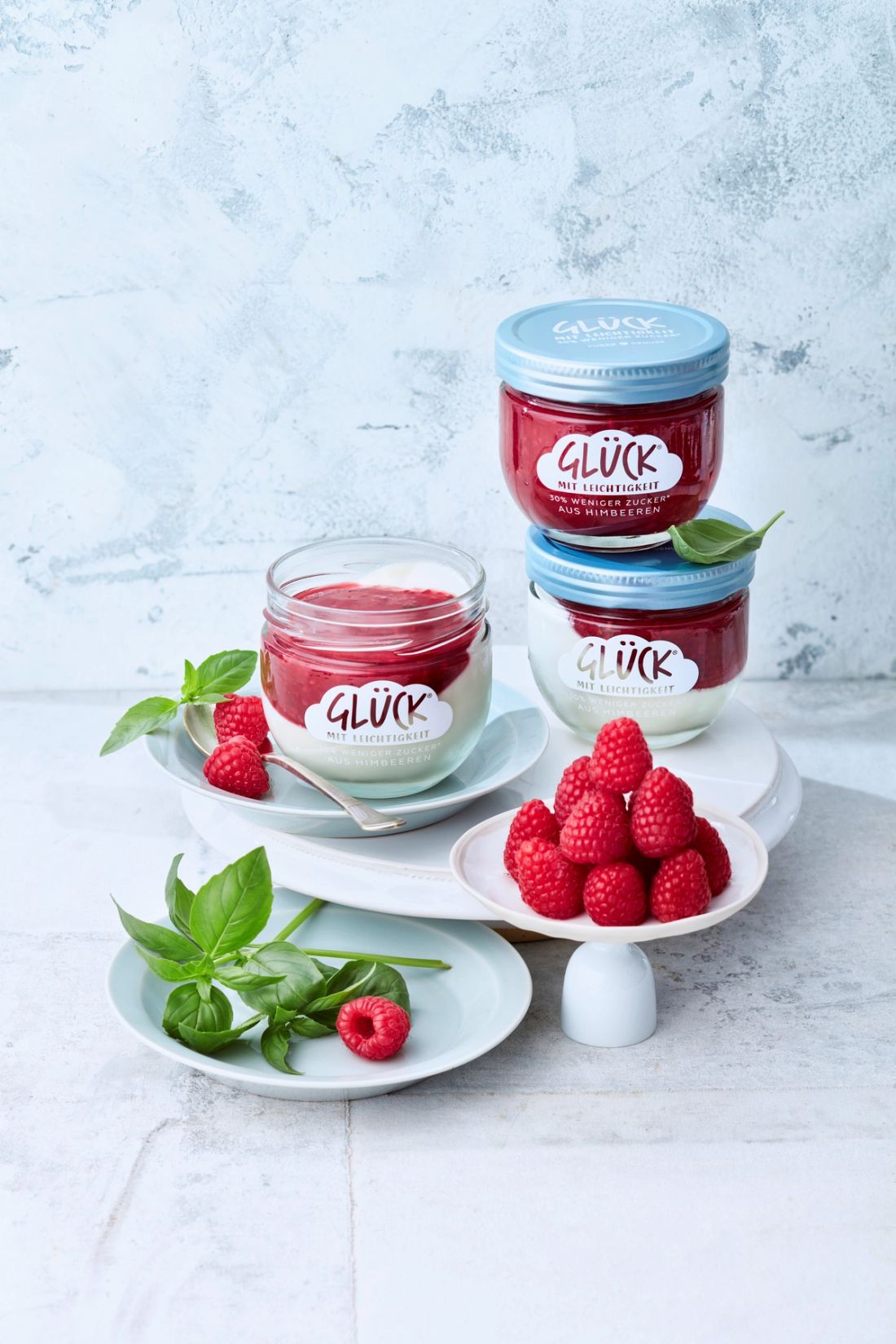 Mandel-Pannacotta mit Himbeersoße im GLÜCK Glas. Daneben Himbeeren und Basilikum. Im Hintergrund ein Glas GLÜCK mit Leichtigkeit aus Himbeeren.