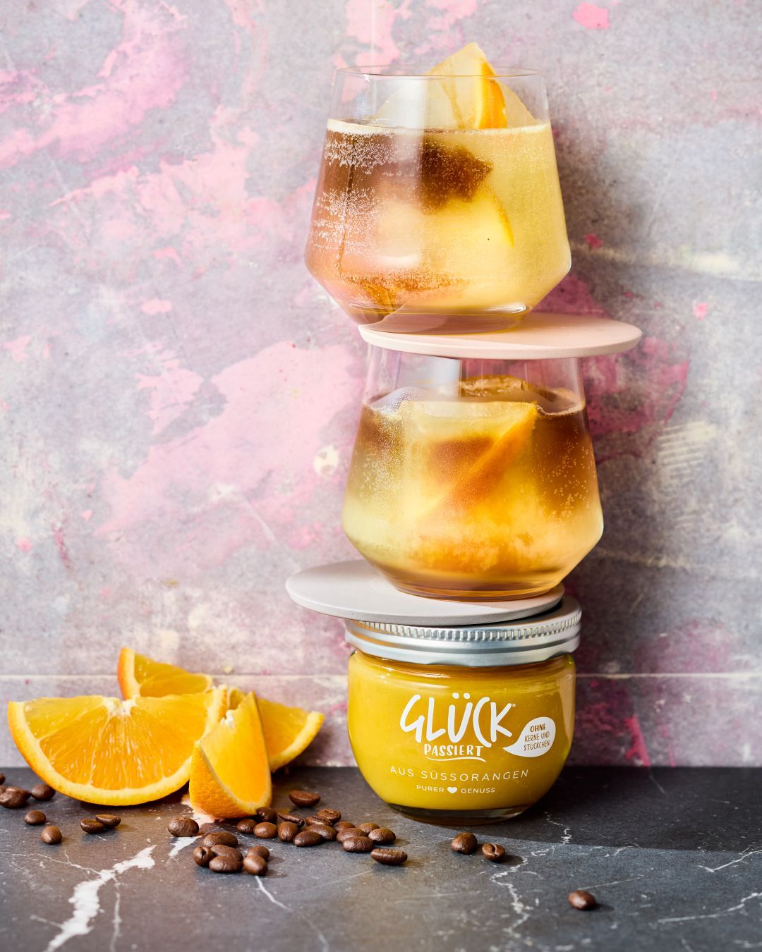 Orangen-Espresso-Tonic mit GLÜCK passiert aus Süßorangen. Es stehen zwei Gläser übereinander auf einem Glas GLÜCK. Daneben liegen Orangenscheiben und Kaffeebohnen.