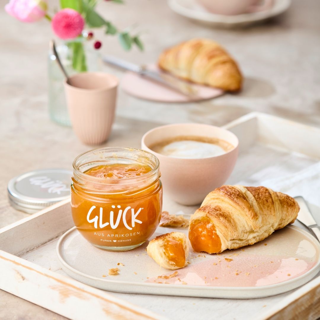 Auf einem rosafarbenen ovalen Teller liegt ein angeschnittenes Croissant gefüllt mit Aprikosenmarmelade, daneben steht ein Glas GLÜCK aus Aprikosen. Im Hintergrund eine Tasse Kaffee, Blumen und ein weiterer Teller mit einem Croissant.