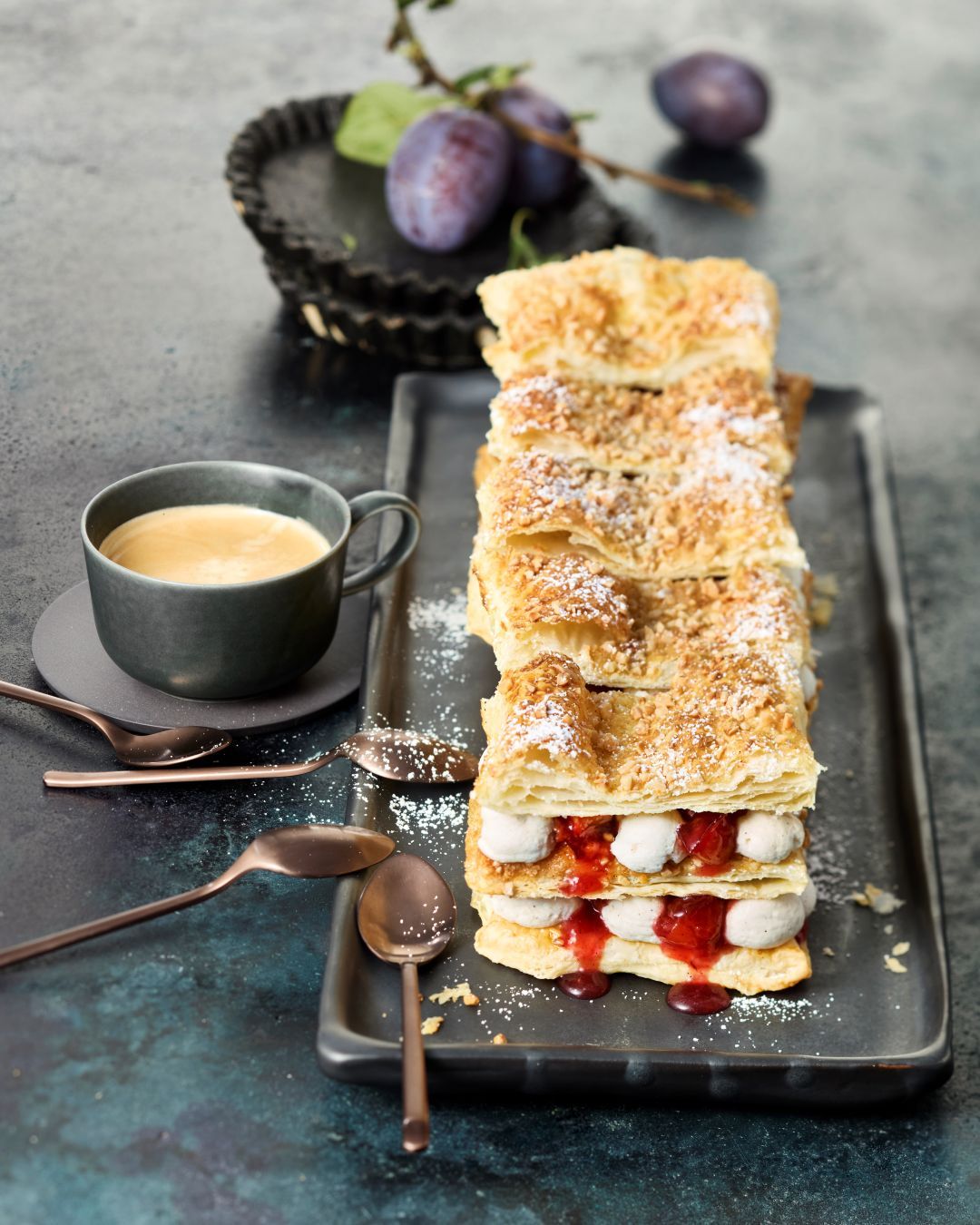 Friesentorte aus Blätterteig, Pflaumen, GLÜCK aus Pflaumen und Sahnecreme. Serviert auf einer schwarzen Platte mit einer Tasse Kaffee.