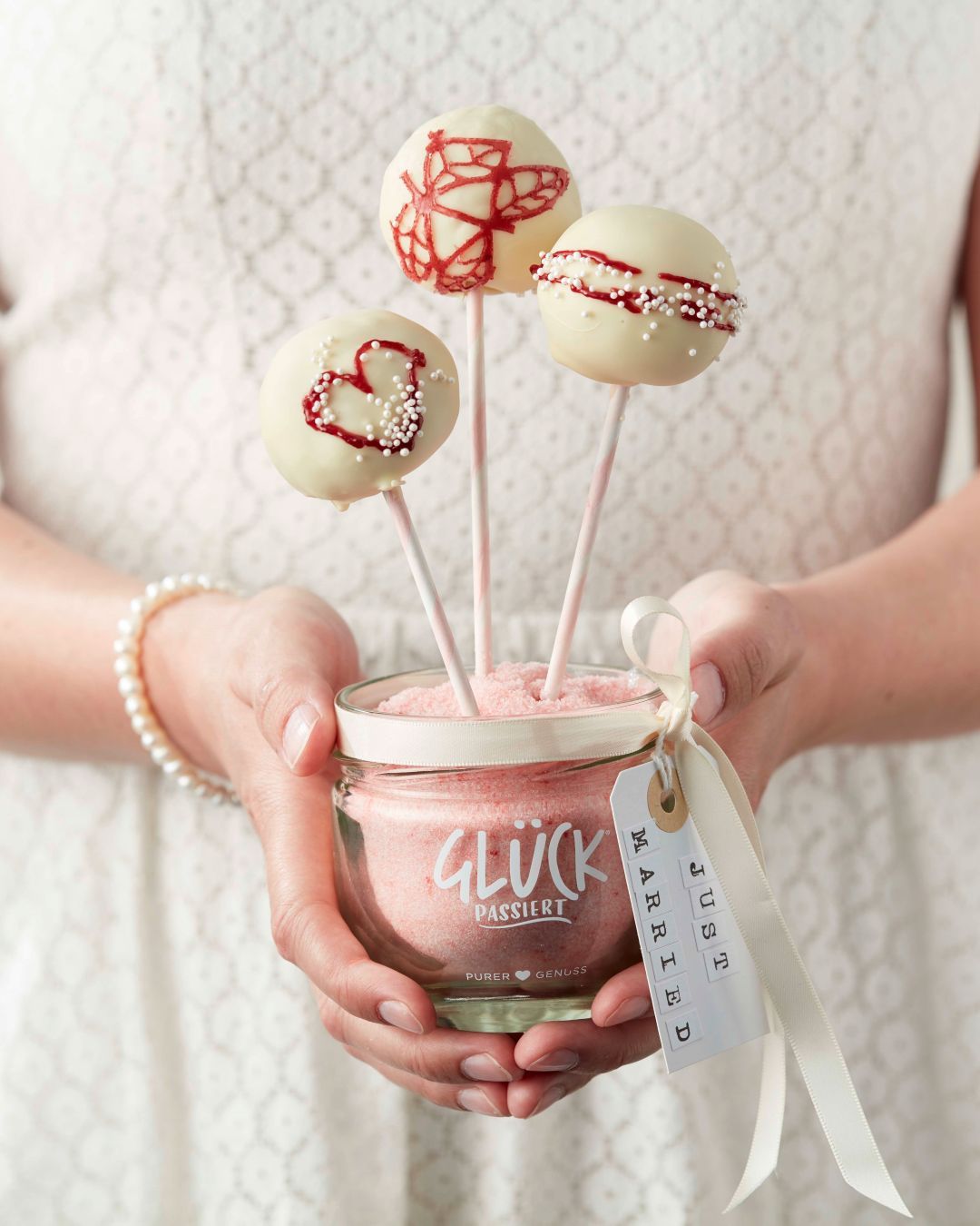Zwei Hände halten ein GLÜCK Glas, das als Halterung für Hochzeitscakepops genutzt wird. Sie sind ummantelt von weißer Schokolade und verziert mit roten Symbolen und weißen Streuseln.