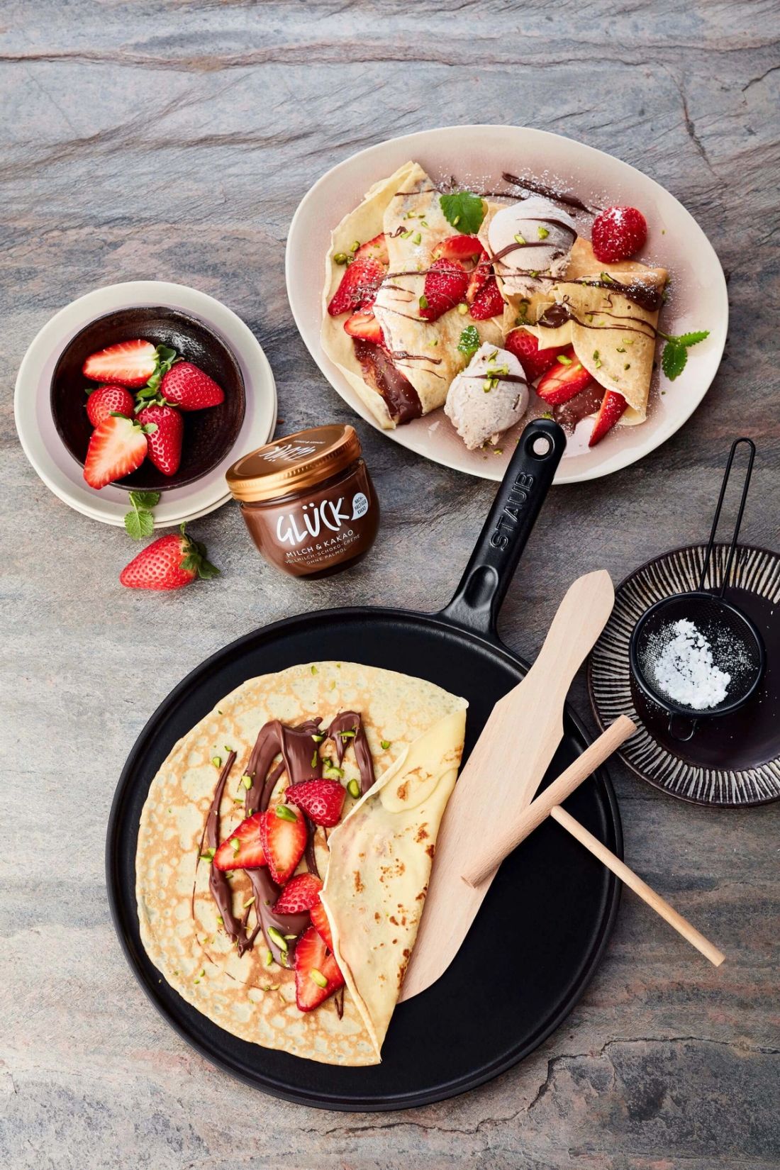 Crepes mit Haselnuss-Nicecream, Schokosauce und Erdbeeren auf einer STAUB Crepepfanne. Daneben mehr Beeren und ein Glas GLÜCK Milch & Kakao.