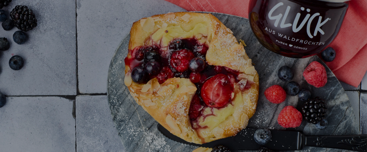 Ein Puddingteilchen mit verschiedenen Beeren und Mandeln auf einer grauen Schieferplatte. Daneben zur Dekoration einige frische Beeren und ein Glas GLÜCK aus Waldfrüchten. Über dem Bild liegt ein leichter Verdunkelungsfilter.