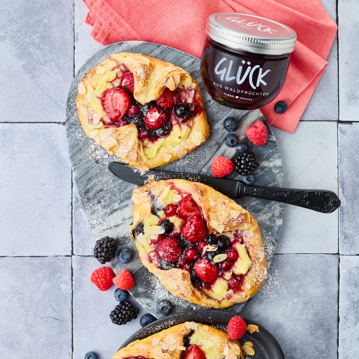 Ein Puddingteilchen mit verschiedenen Beeren und Mandeln auf einer grauen Schieferplatte. Daneben zur Dekoration einige frische Beeren und ein Glas GLÜCK aus Waldfrüchten.