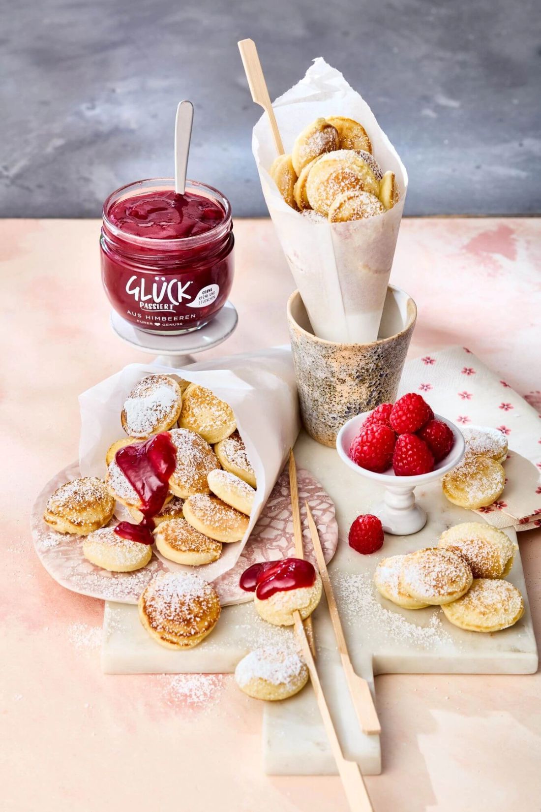 Poffertjes mit Himbeerpüree in einer Tüte. Daneben ein Glas GLÜCK passiert aus Himbeeren.