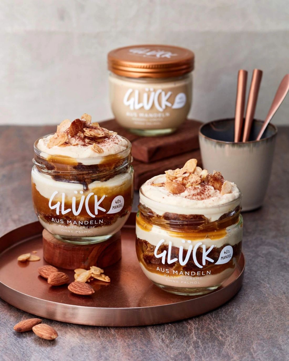 Nuss-Tiramisù im GLÜCK aus Mandeln Glas. Geschichtet sind Löffelbiskuits, Mascarponeceme, und GLÜCK aus Aprikosen. Im Hintergrund ein Glas GLÜCK aus Mandeln. Zur Dekoration liegen Mandeln neben den Gläsern.