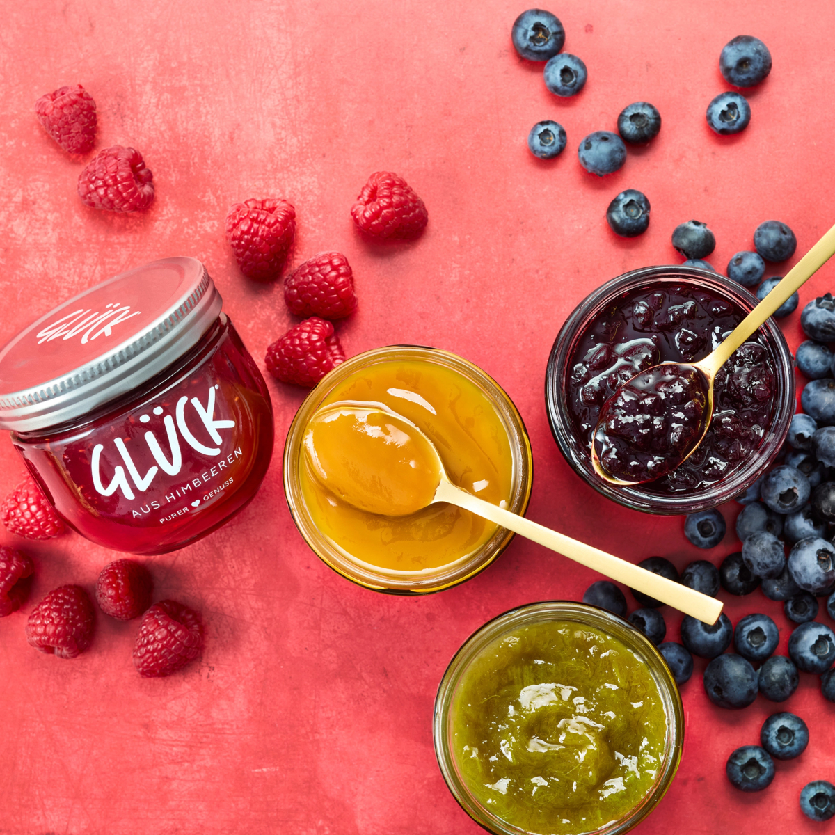 Drei offene Gläser Marmelade. Ein GLÜCK aus Rhabarber, ein GLÜCK aus Heidelbeeren und ein GLÜCK passiert aus Mango. In zwei Gläsern liegen Löffel. Daneben liegt ein Glas GLÜCK aus Himbeeren. Darum verteilt liegen einige frische Beeren auf rosarotem Hintergrund.