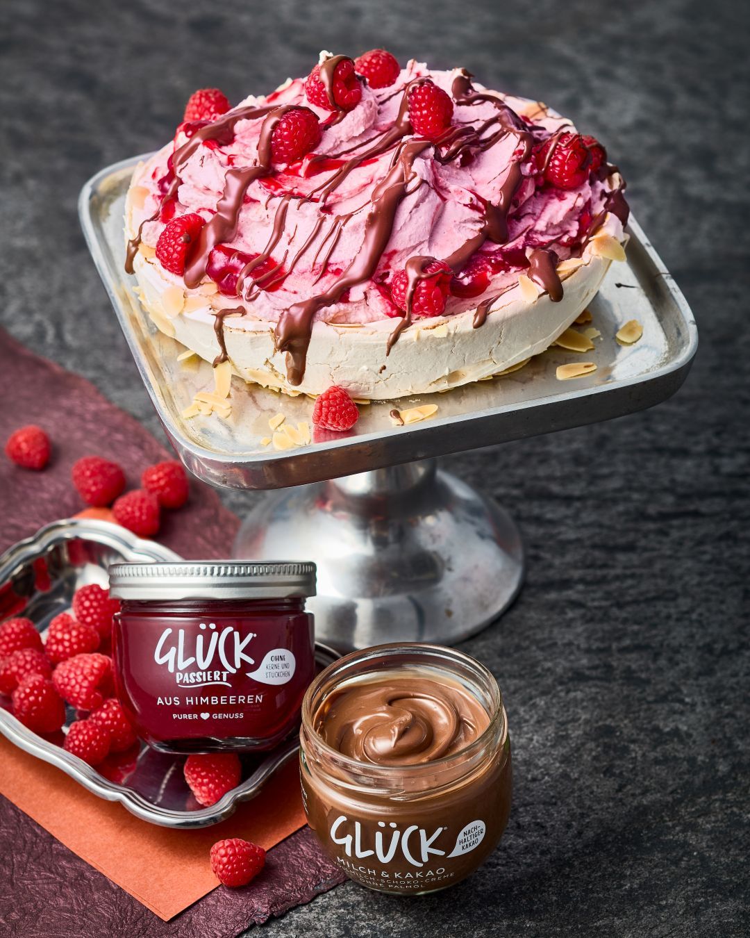 Mandelbaiser-Pavlova auf einer Servierplatte, angerichtet mit Himbeeren, GLÜCK passiert aus Himbeeren und GLÜCK Milch & Kakao.