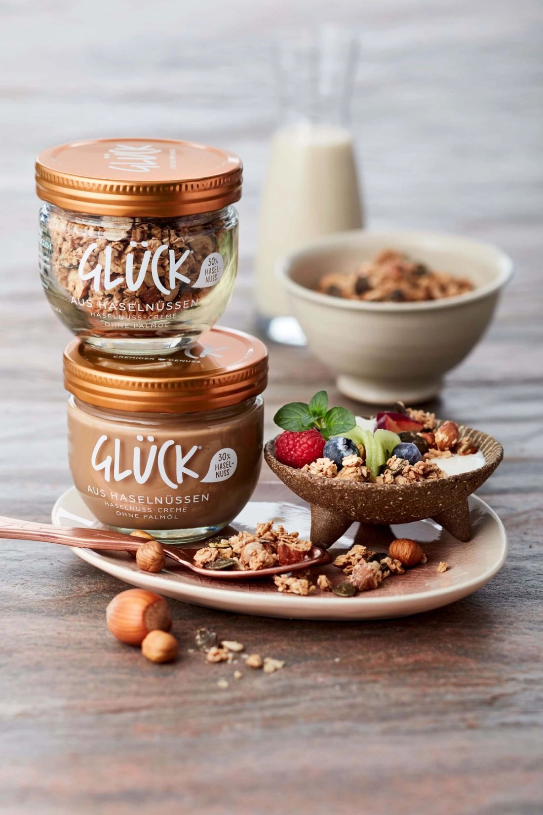 Haselnuss-Crunchy-Müsli im GLÜCK Glas. Darunter ein Glas GLÜCK aus Haselnüssen. Beeren und Nüsse zum Garnieren.