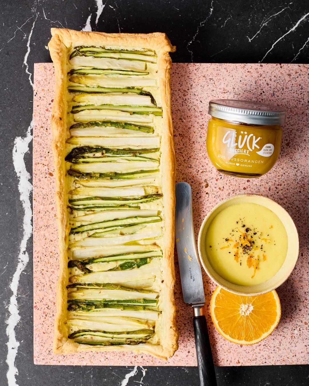 Spargel-Tarte mit Hollandaise mit GLÜCK passiert aus Süßorangen. Daneben steht ein Glas der Hollandaise. Dabei liegen eine halbe Orange und ein Glas GLÜCK passiert aus Süßorangen.