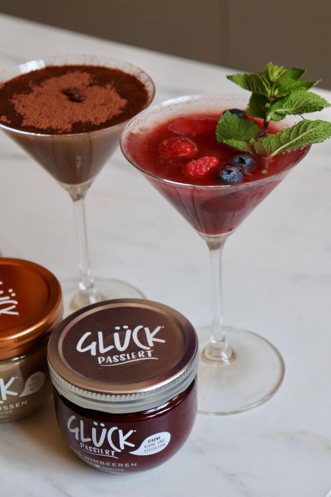 Zwei Cocktails, garniert mit Beeren und Minze sowie Kakaopulver. Daneben ein Glas GLÜCK passiert.