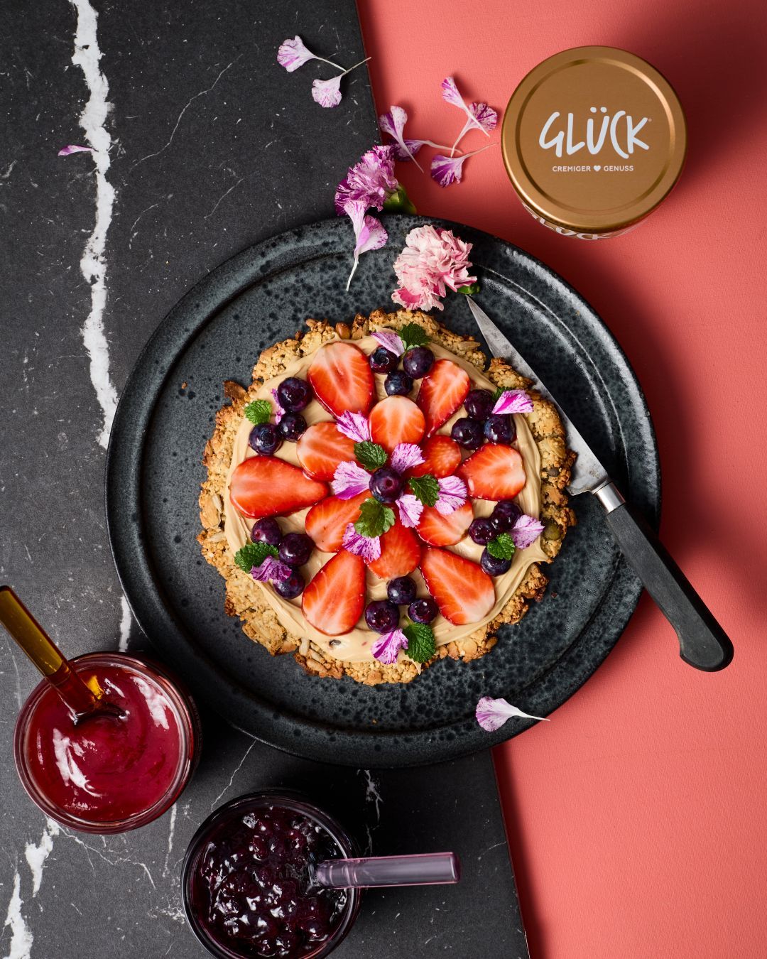 Granola Pizza, belegt mit GLÜCK aus Mandeln sowie frischen Beeren. Zum Garnieren essbare Blüten und Minze.