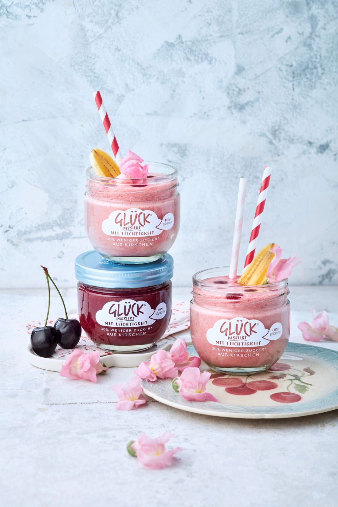 Kirsch-Bananen-Milkshake im Glas von GLÜCK passiert mit Leichtigkeit aus Kirschen. Zur Dekoration Blütenblätter und Kirschen auf einem Porzellanteller mit Kirschprint.