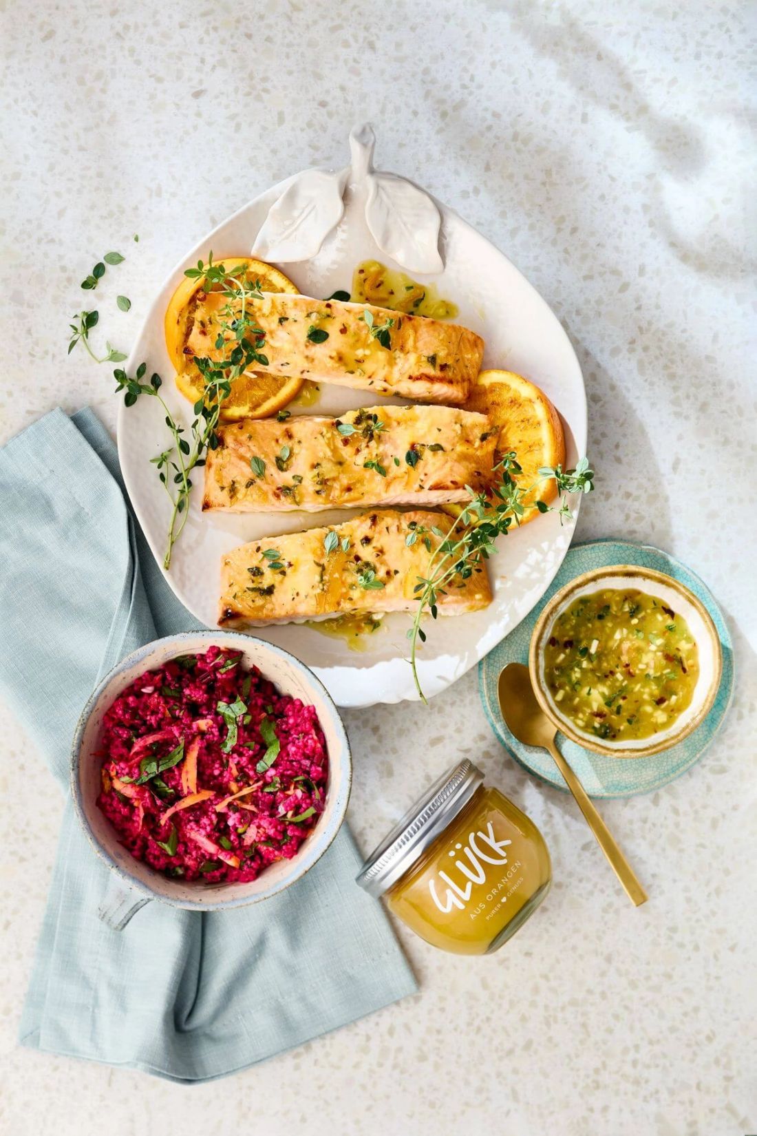 Gegrillter Orangenlachs auf einem Teller mit Sauce und Couscoussalat. Daneben ein Glas GLÜCK aus Orangen.
