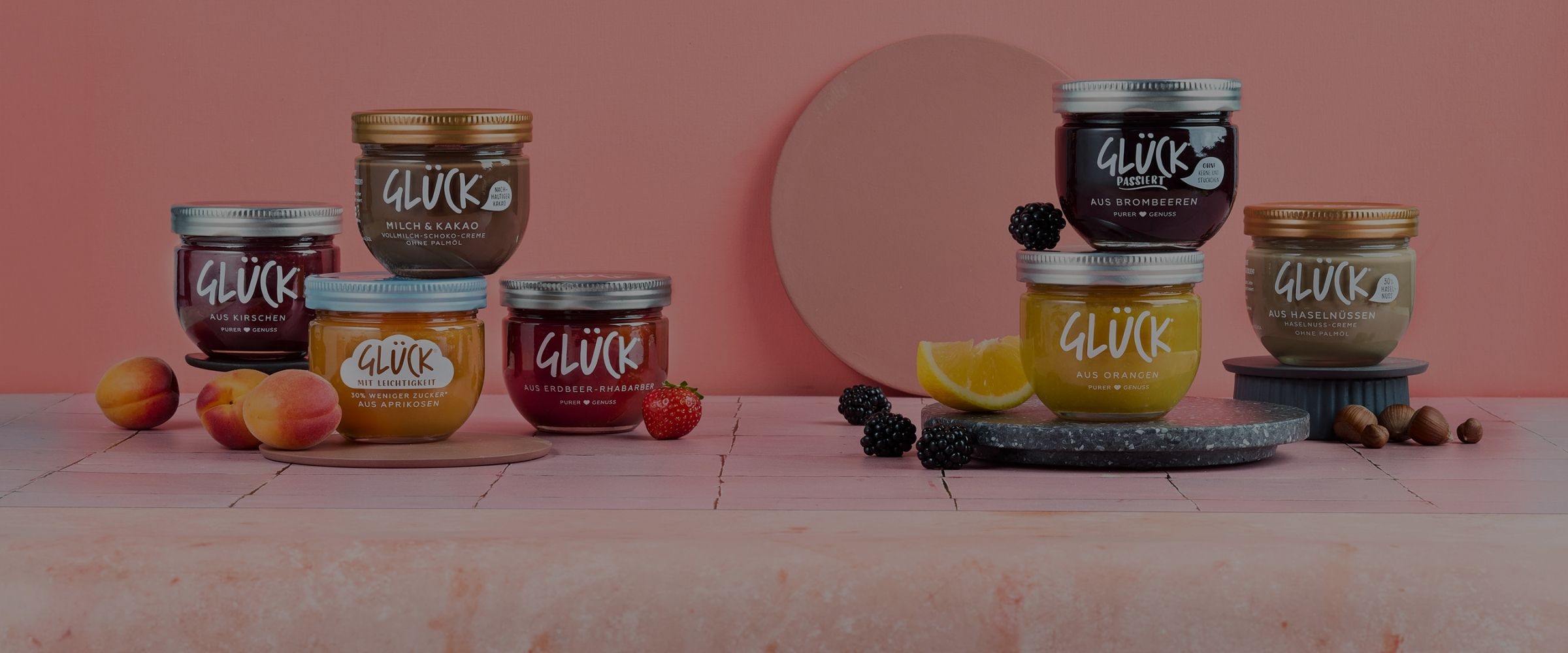 Viele Marmeladengläser stehen auf rosafarbenen Fliesen. Links im Bild stehen vier Sorten GLÜCK, daneben liegen Aprikosen und Erdbeeren. Auf der rechten Seite stehen drei Gläser GLÜCK Produkte, daneben liegen Orangenscheiben, Brombeeren und Haselnüsse.
