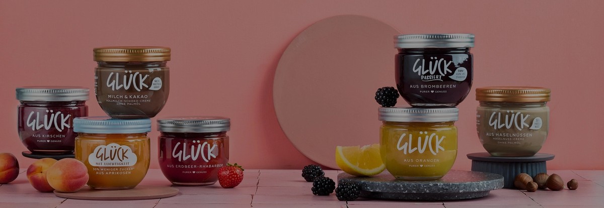 Viele Marmeladengläser stehen auf rosafarbenen Fliesen. Links im Bild stehen vier Sorten GLÜCK, daneben liegen Aprikosen und Erdbeeren. Auf der rechten Seite stehen drei Gläser GLÜCK Produkte, daneben liegen Orangenscheiben, Brombeeren und Haselnüsse.