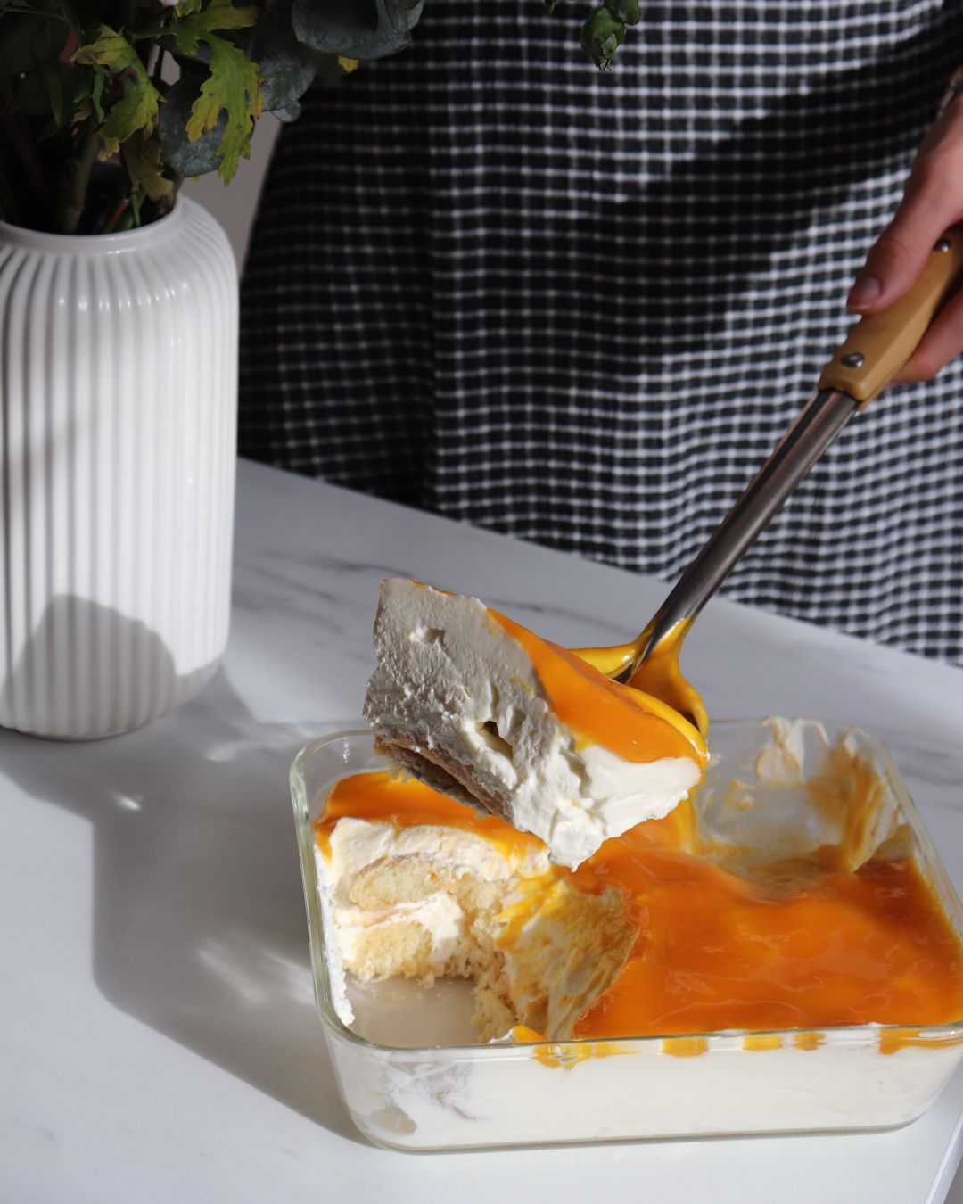 Mango-Tiramisu in einer Form, es wird mit einer Kelle ein Stück angehoben.