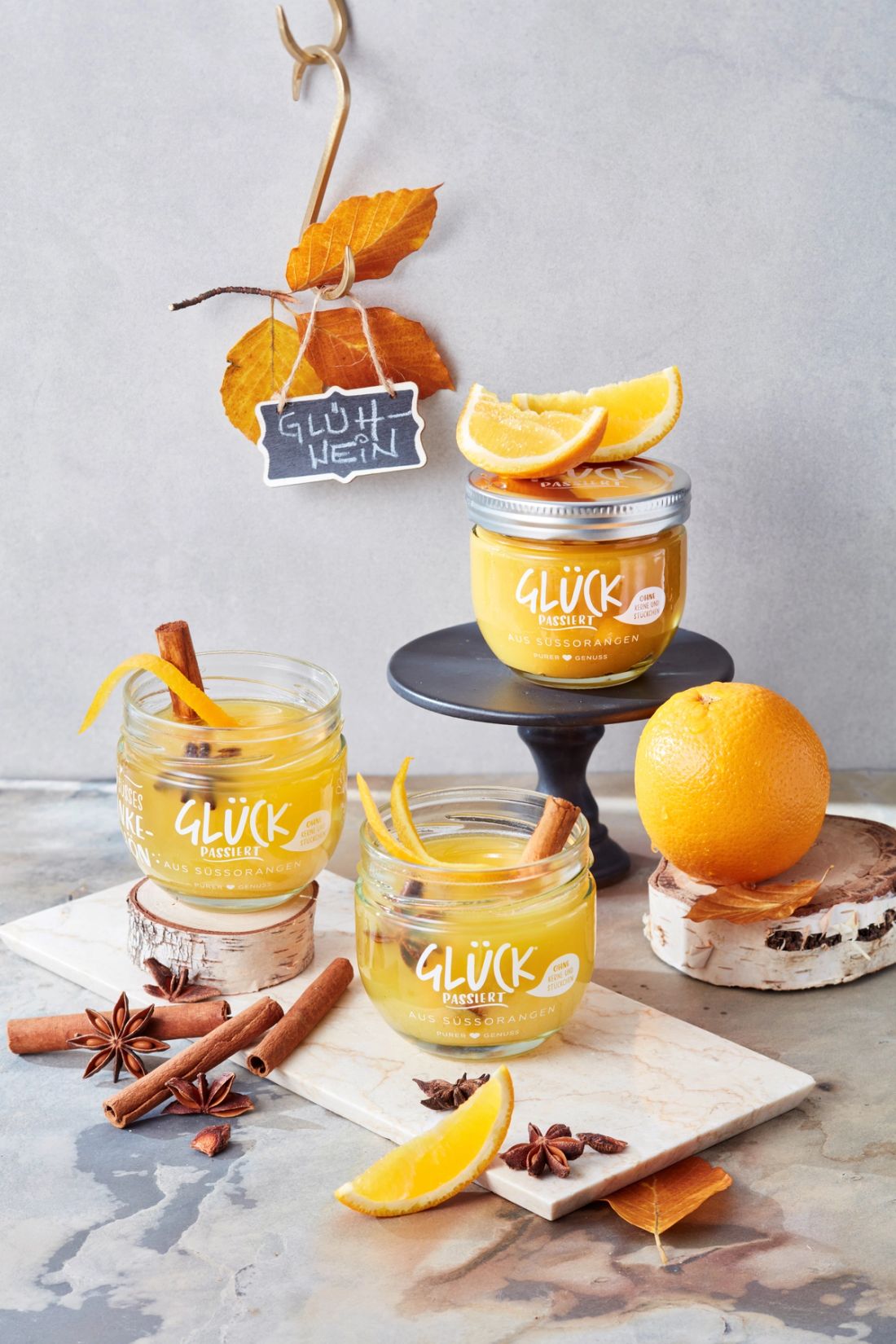 Drei Marmeladengläser gefüllt mit Orangen-Glühwein. Darum winterlich-herbstliche Deko. Im Hintergrund steht ein Glas GLÜCK passiert aus Süßorangen.
