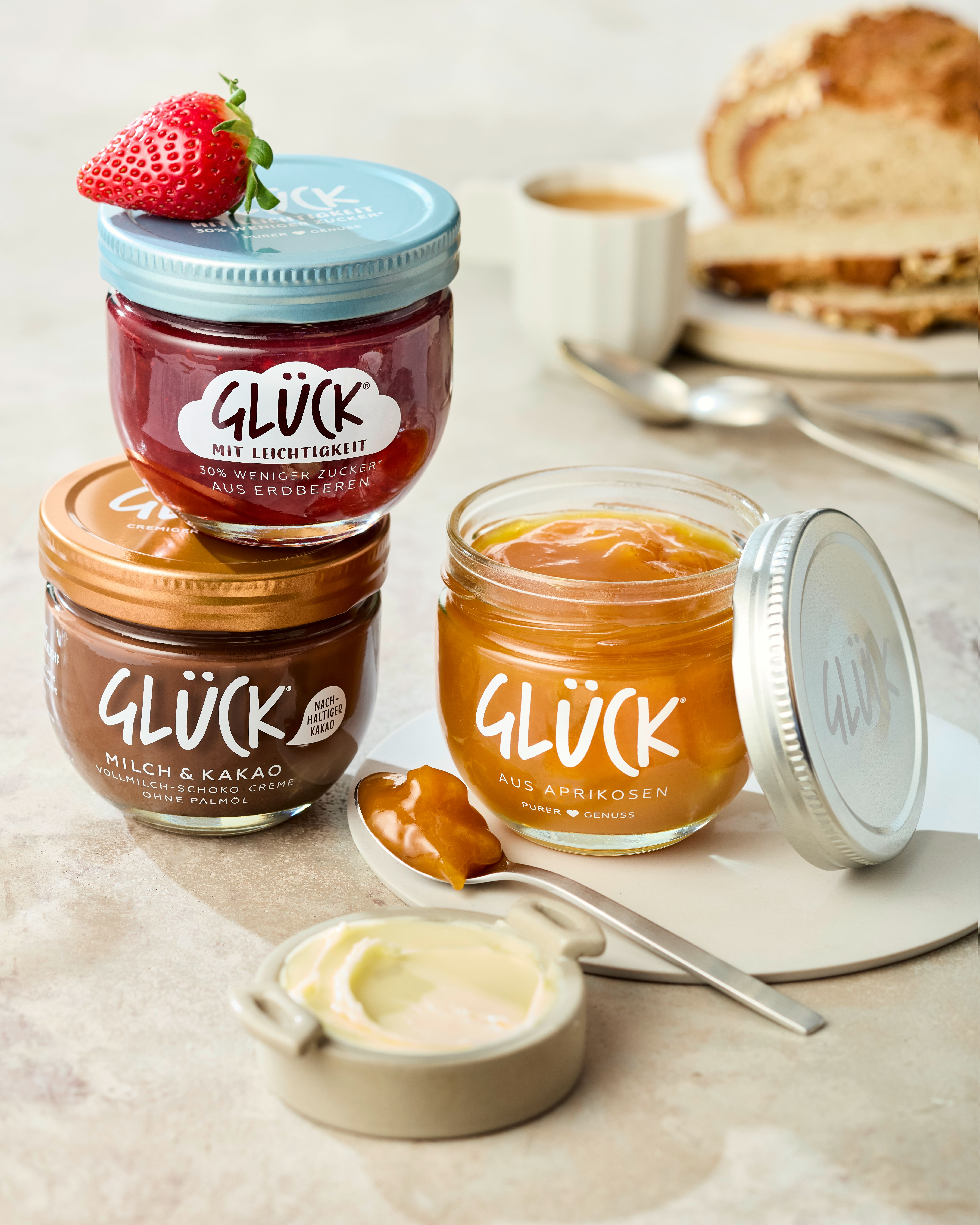 Rechts steht ein offenes Glas GLÜCK aus Aprikosen, davor liegt ein Löffel mit etwas Marmelade drauf. Links steht ein Glas GLÜCK Milch & Kakao, darüber steht GLÜCK mit Leichtigkeit aus Erdbeeren. Im Hintergrund sieht man Brot und andere Frühstücksutensilien.