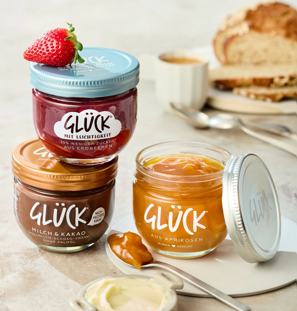 Rechts steht ein offenes Glas GLÜCK aus Aprikosen, davor liegt ein Löffel mit etwas Marmelade drauf. Links steht ein Glas GLÜCK Milch & Kakao, darüber steht GLÜCK mit Leichtigkeit aus Erdbeeren. Im Hintergrund sieht man Brot und andere Frühstücksutensilien.