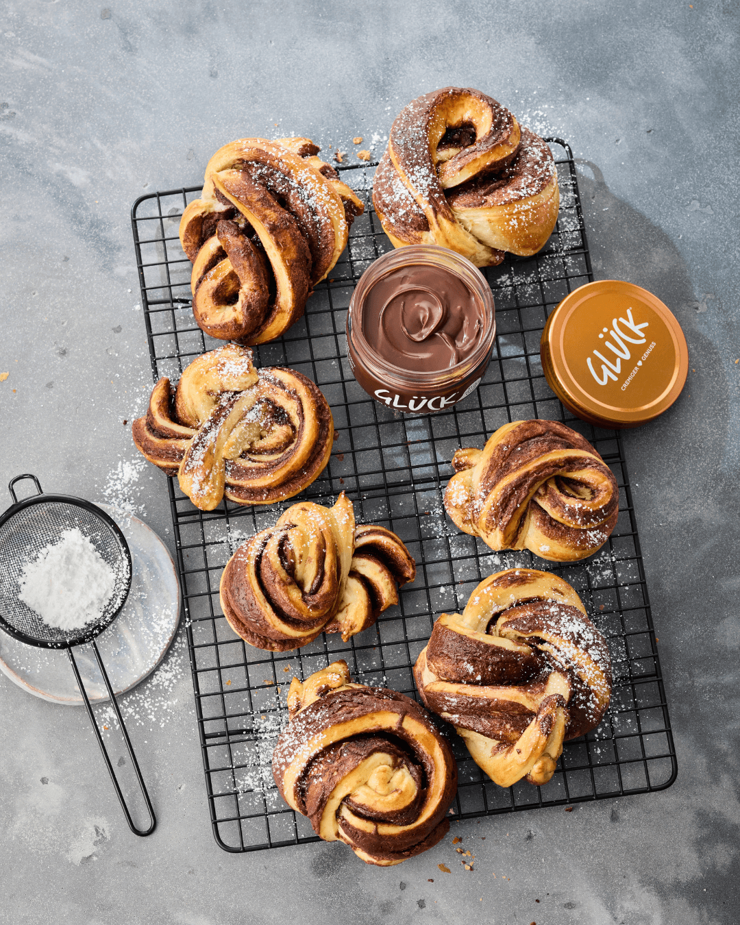 Kakao Kardamom Buns auf einem Gitter. Daneben Puderzucker und ein Glas GLÜCK.