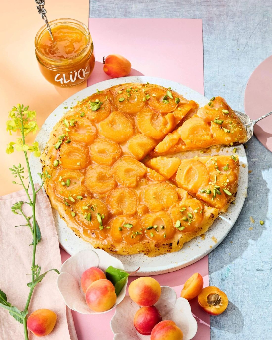 Aprikosen Tarte Tatin, garniert mit Pistazien. Daneben Aprikosen und ein Glas GLÜCK aus Aprikosen.