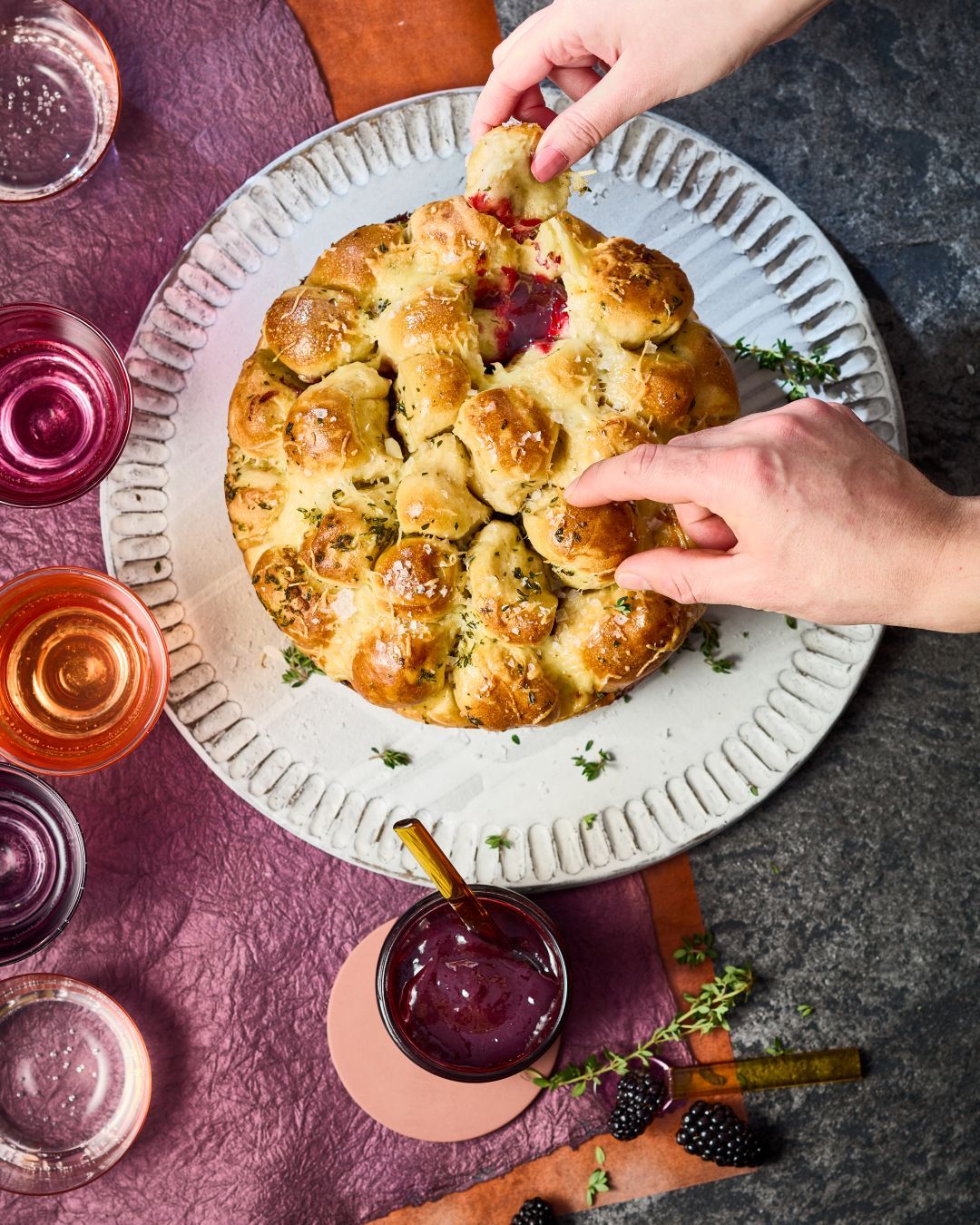 Laugen-Monkey-Bread wird von mehreren Händen auseinandergezogen. Daneben steht ein Glas GLÜCK passiert aus Brombeeren.