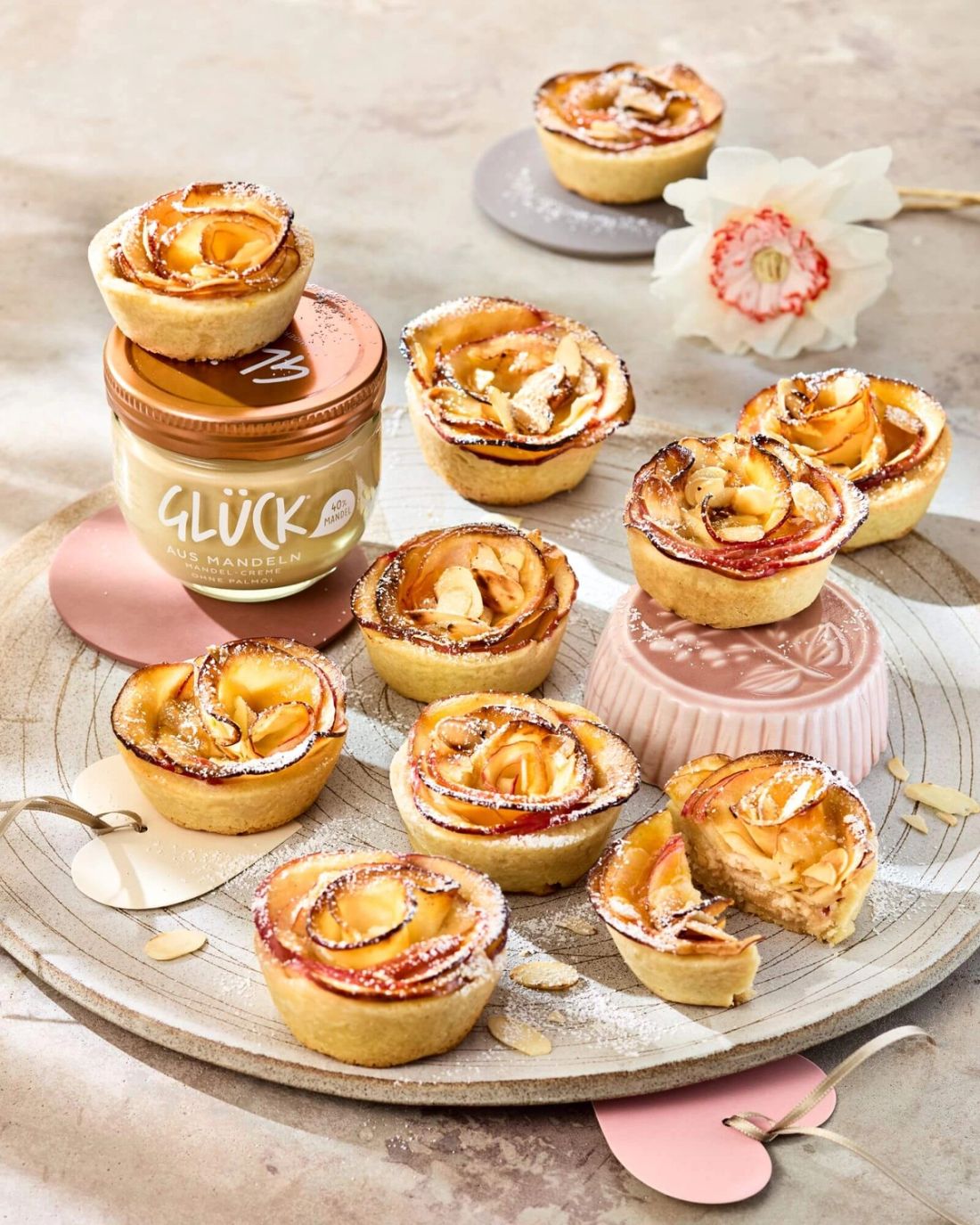 Mandeltörtchen mit Apfelrosen garniert mit Puderzucker. Daneben ein Glas GLÜCK aus Mandeln.