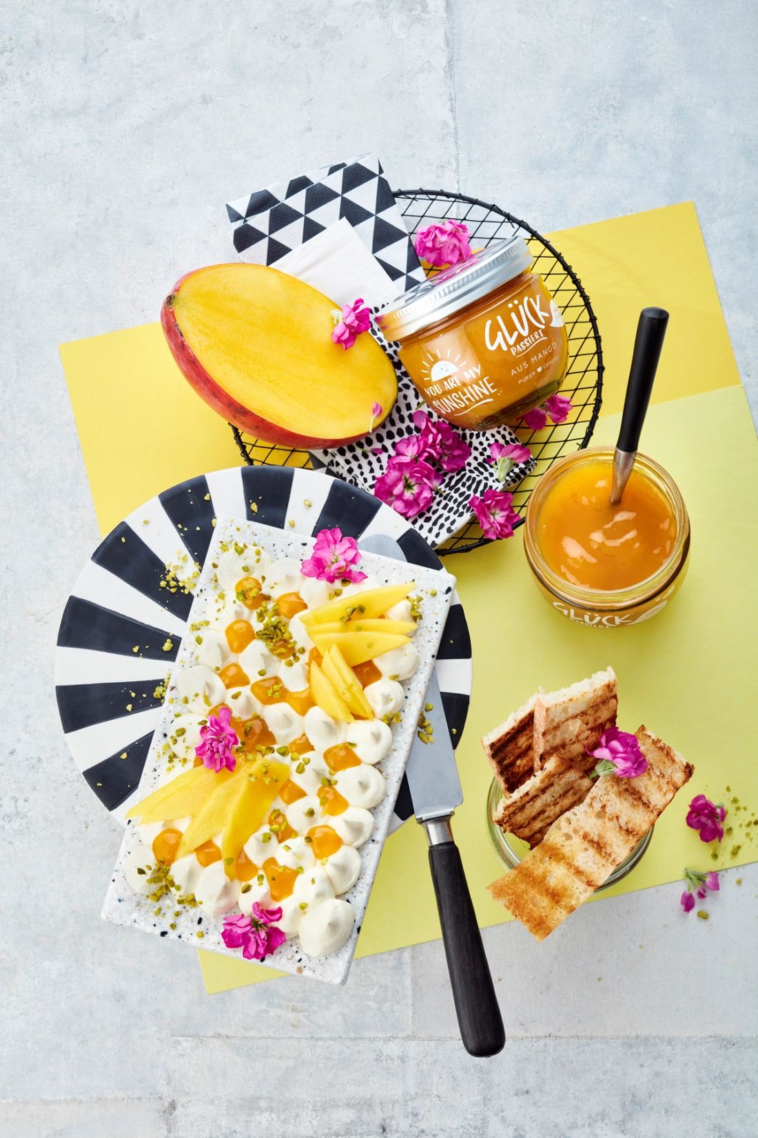 Süßes Butterboard mit Mangomarmelade, garniert mit Mango und essbaren Blüten. Daneben Brot und GLÜCK passiert aus Mango.