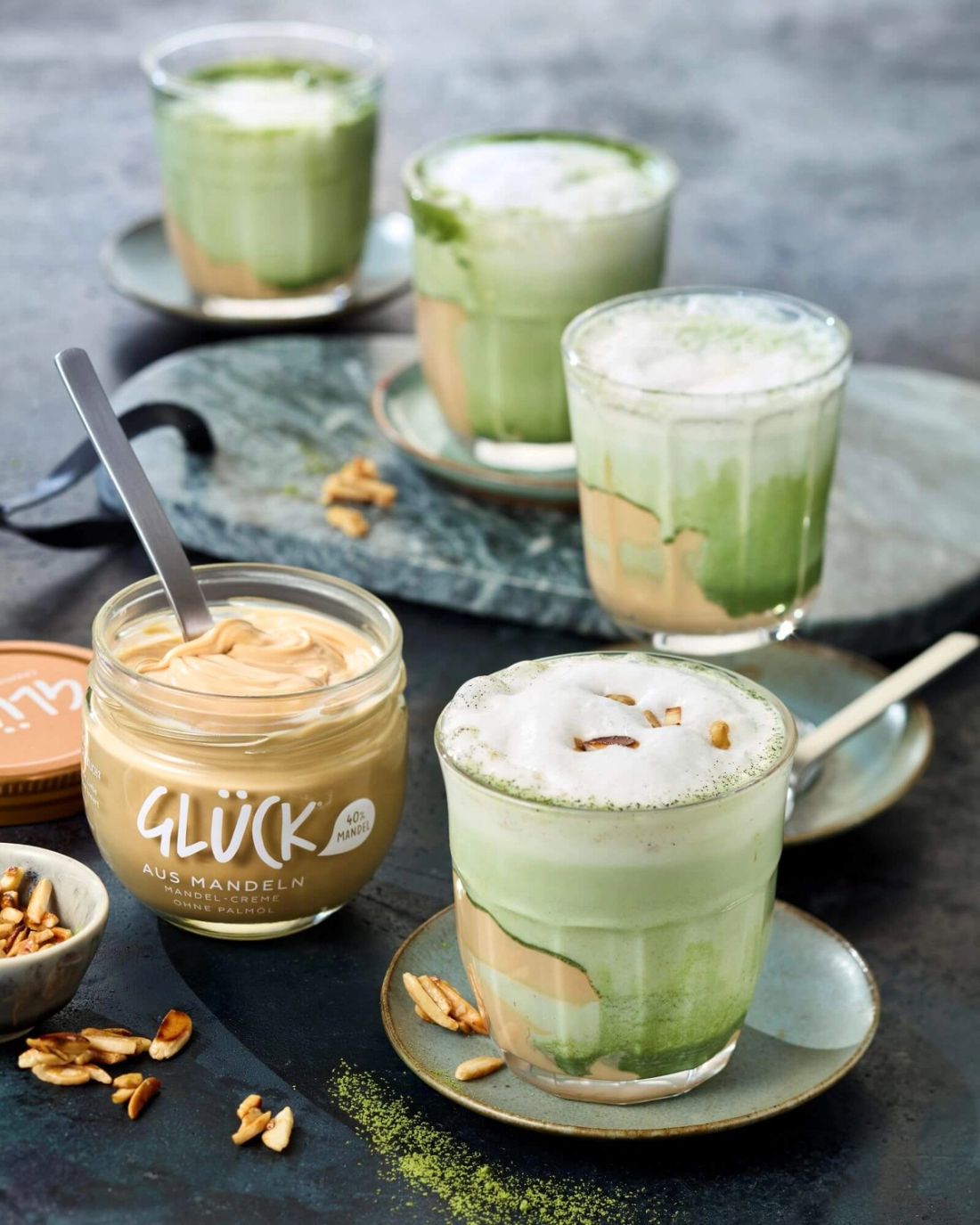 Vier Mandel-Matcha-Latte hintereinander. Daneben ein Glas GLÜCK aus Mandeln.