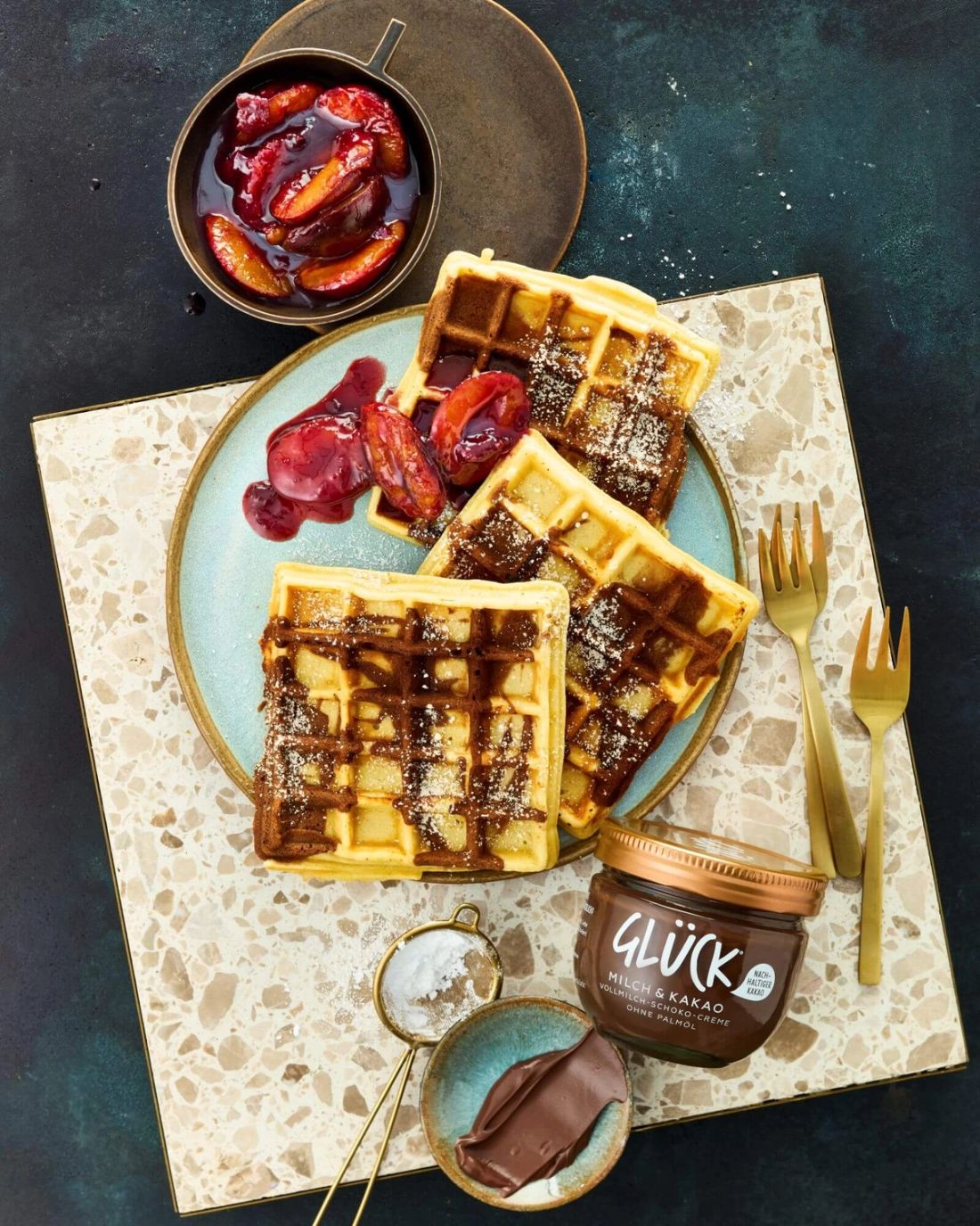 Waffeln mit Schoko-Marmorierung auf einem Teller. Daneben Pflaumensauce, ein Glas GLÜCK Milch & Kakao und Besteck